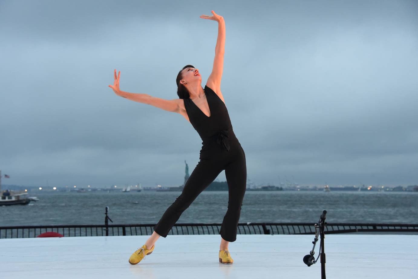 In a black jumpsuit with a deep V-neck and yellow tap shoes, Demi Remick points one foot to the side as her arms are lifted in a V, wrists flexed.